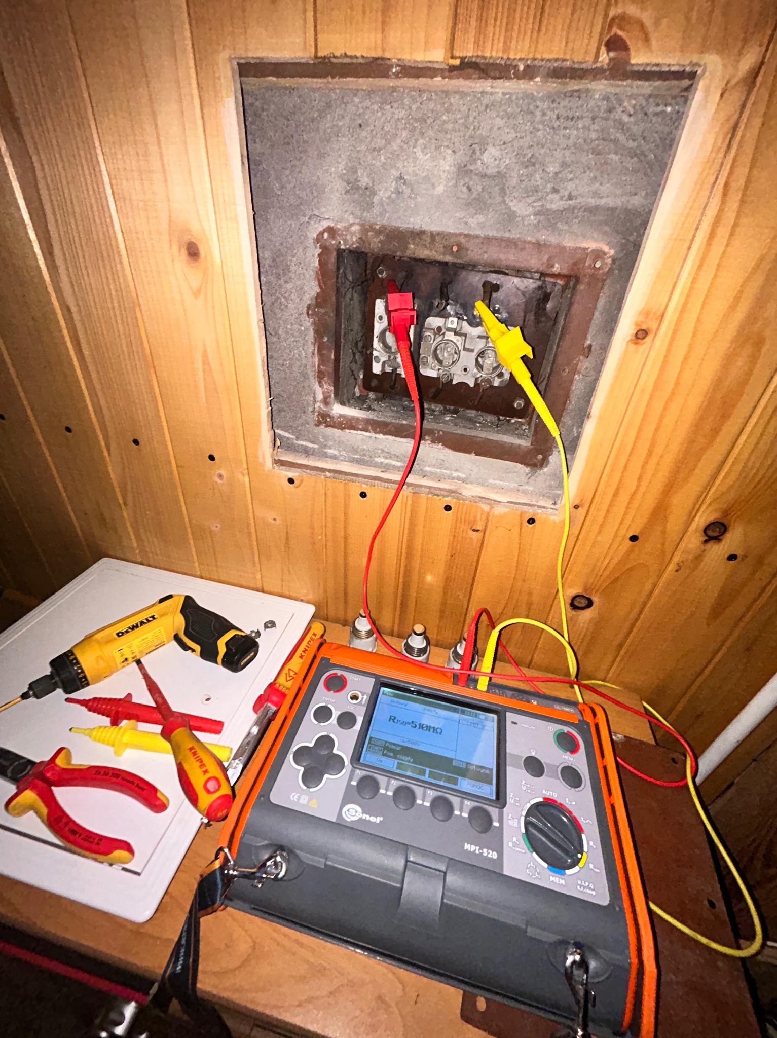 Pomiar rezystancji izolacji w skrzynce z bezpiecznikami, przy użyciu miernika Sonel MPI-520 z podłączonymi przewodami pomiarowymi zakończonymi krokodylkami; obok leżą narzędzia: wkrętak DeWalt...