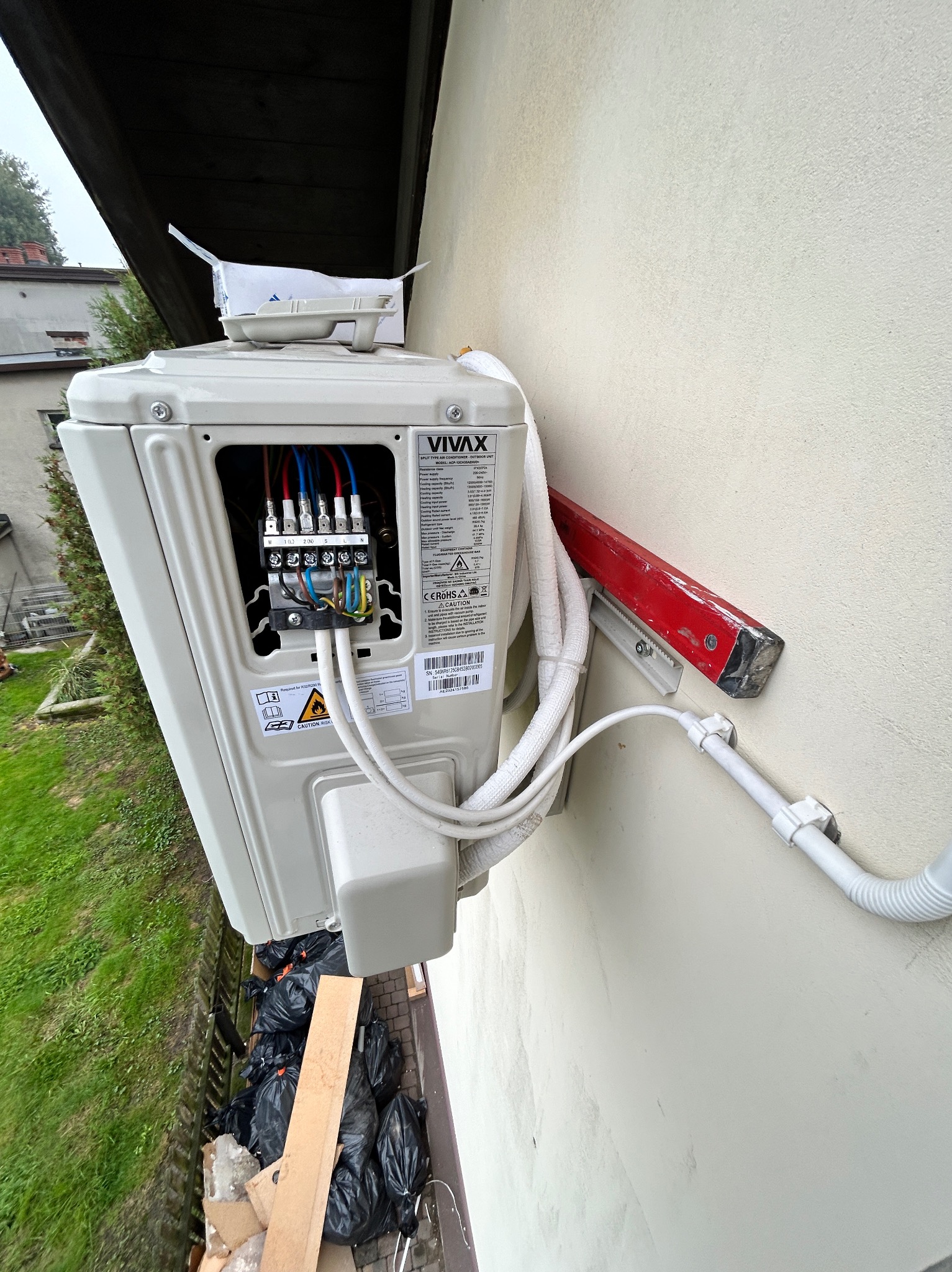 Zewnętrzna jednostka klimatyzacji marki Vivax zamontowana na elewacji budynku, widoczne podłączenia elektryczne oraz rury chłodnicze, poziomica przytwierdzona do ściany, śmieci w czarnych workach...