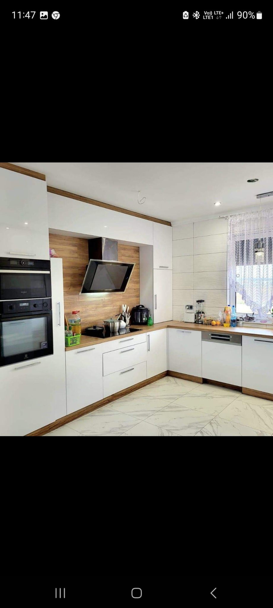 Nowoczesna kuchnia z białymi szafkami, drewnianymi akcentami i marmurową podłogą; na blacie widoczne akcesoria kuchenne i okno z koronkową firanką.