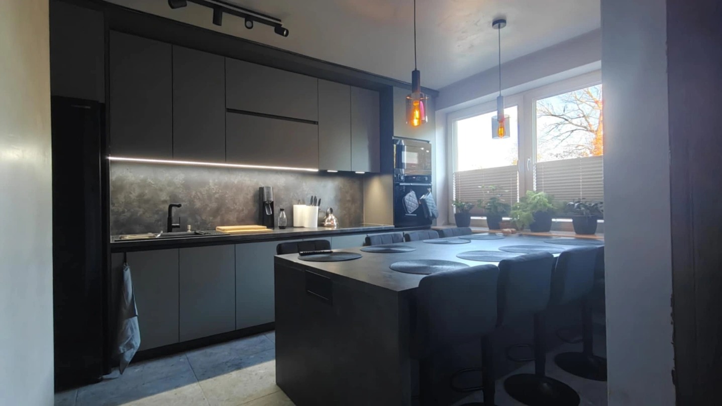 Nowoczesna, szara kuchnia z wyspą i oświetleniem LED pod szafkami, widok zza filaru.