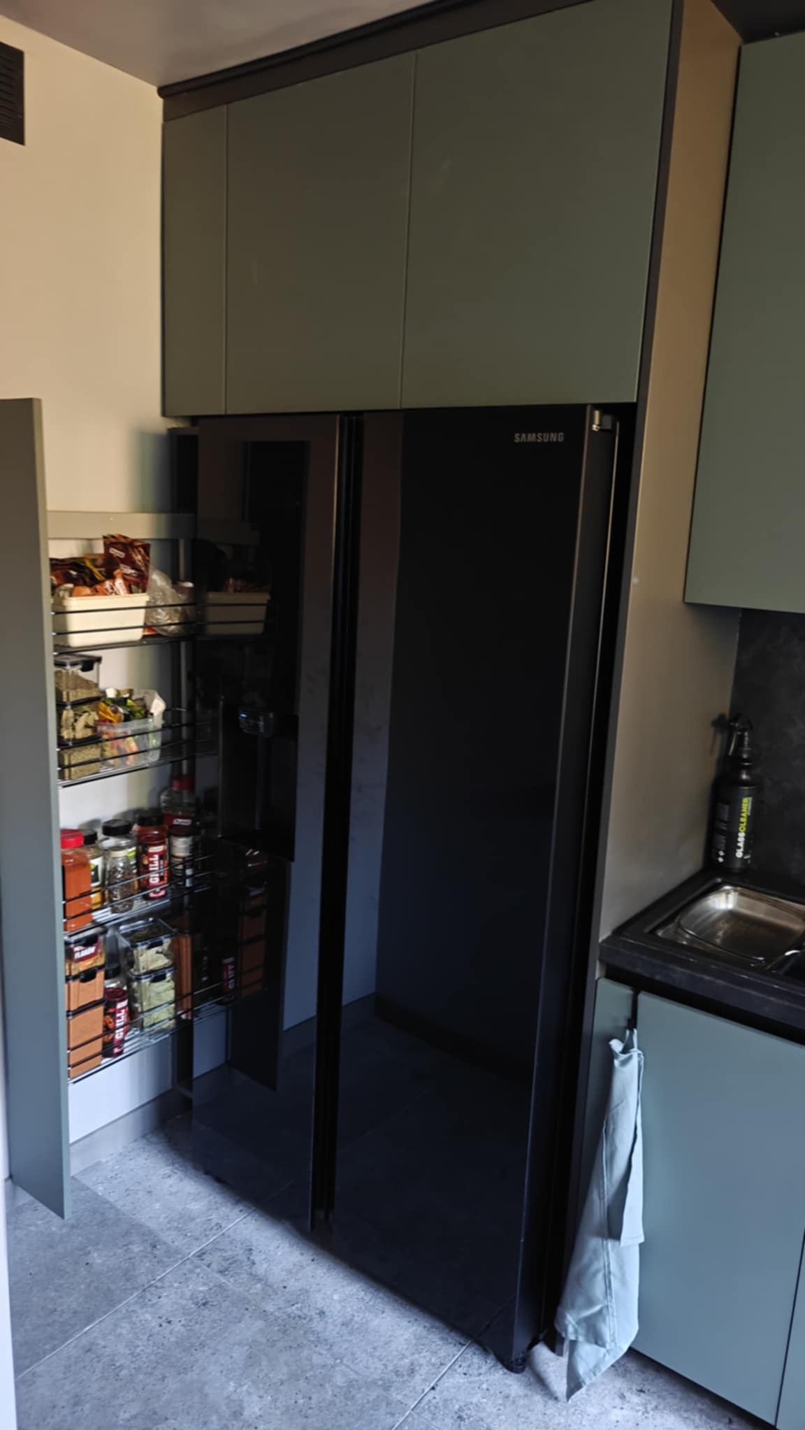 Nowoczesna kuchnia z szarymi szafkami, czarną lodówką Samsung side-by-side i wysuwaną spiżarnią z metalowymi koszami wypełnionymi produktami spożywczymi.