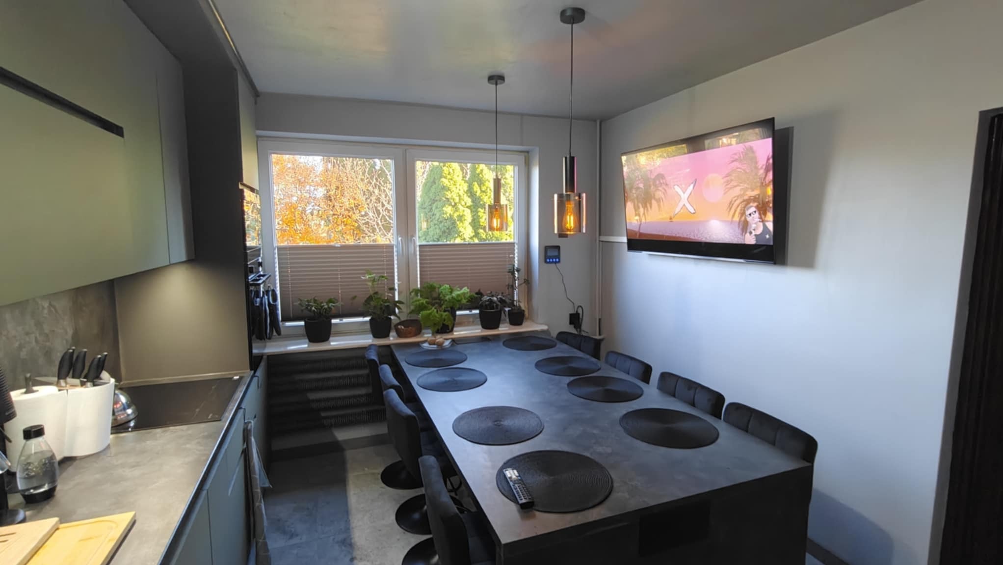 Nowoczesna kuchnia z szarymi szafkami, blatem i dużym stołem jadalnym z czarnymi podkładkami, oświetlona wiszącymi lampami z bursztynowymi kloszami, z telewizorem na ścianie.