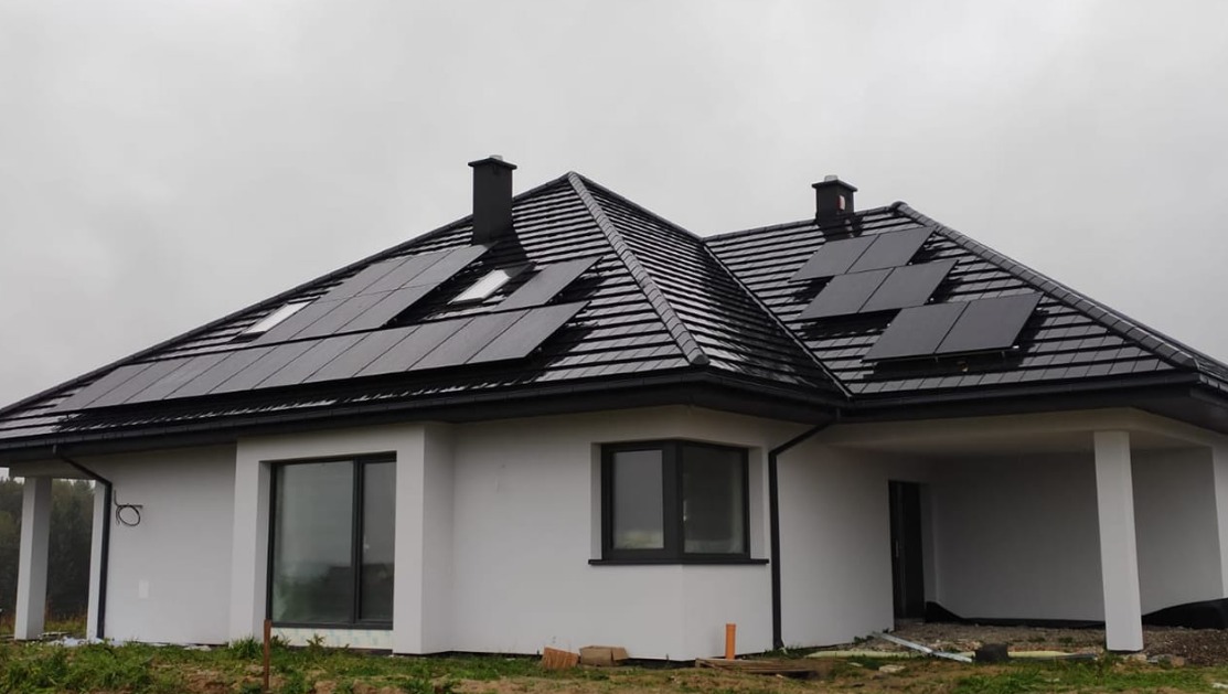 Nowoczesny dom jednorodzinny z ciemną dachówką i panelami słonecznymi na dachu, widoczne dwa kominy i okno dachowe, pochmurne niebo.