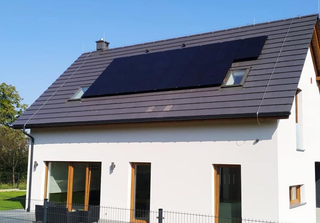 Dom jednorodzinny z szarym dachem pokrytym panelami fotowoltaicznymi, widoczne okna dachowe i jasna elewacja budynku.