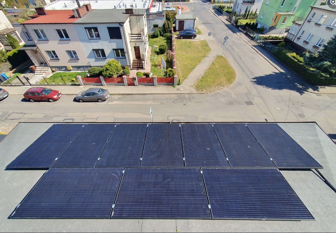 Widok z góry na panele słoneczne na płaskim dachu budynku mieszkalnego w słoneczny dzień; w tle ulica z samochodami i zabudowa jednorodzinna.