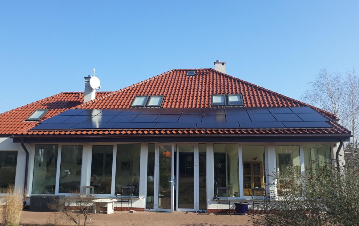Dom jednorodzinny z czerwonym dachem krytym dachówką i zamontowanymi panelami słonecznymi; widoczne okna dachowe, antena satelitarna, duże okna tarasowe.