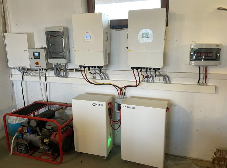 Dwa białe magazyny energii Power Lab na kółkach, podłączone kablami do dwóch falowników na ścianie. Obok agregat prądotwórczy Honda w czerwonej ramie, a także skrzynki elektryczne i bezpieczniki.