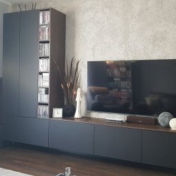 Era meble Bogumił Woźniak - Nowoczesny salon z zabudową meblową w kolorze grafitowym, obejmującą szafę z półkami na płyty, szafkę RTV oraz dekoracje: zegar, figurka anioła i suszone trawy. Na ścianie widoczna faktura...