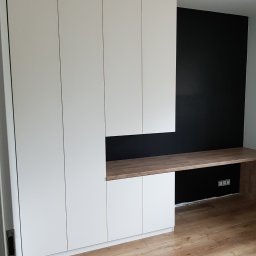 Era meble Bogumił Woźniak - Biała zabudowa meblowa z blatem roboczym w kolorze drewna, na tle czarnej ściany, widoczne gniazdka elektryczne.