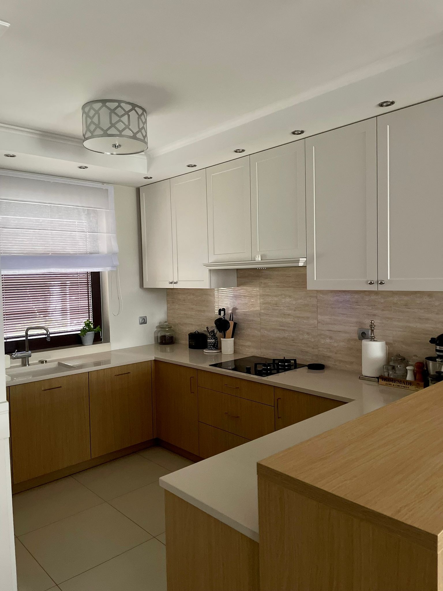 Jasna kuchnia z drewnianymi dolnymi szafkami i białymi górnymi, blat z jasnego kamienia, okno z roletami rzymskimi, widoczna płyta gazowa i akcesoria kuchenne.