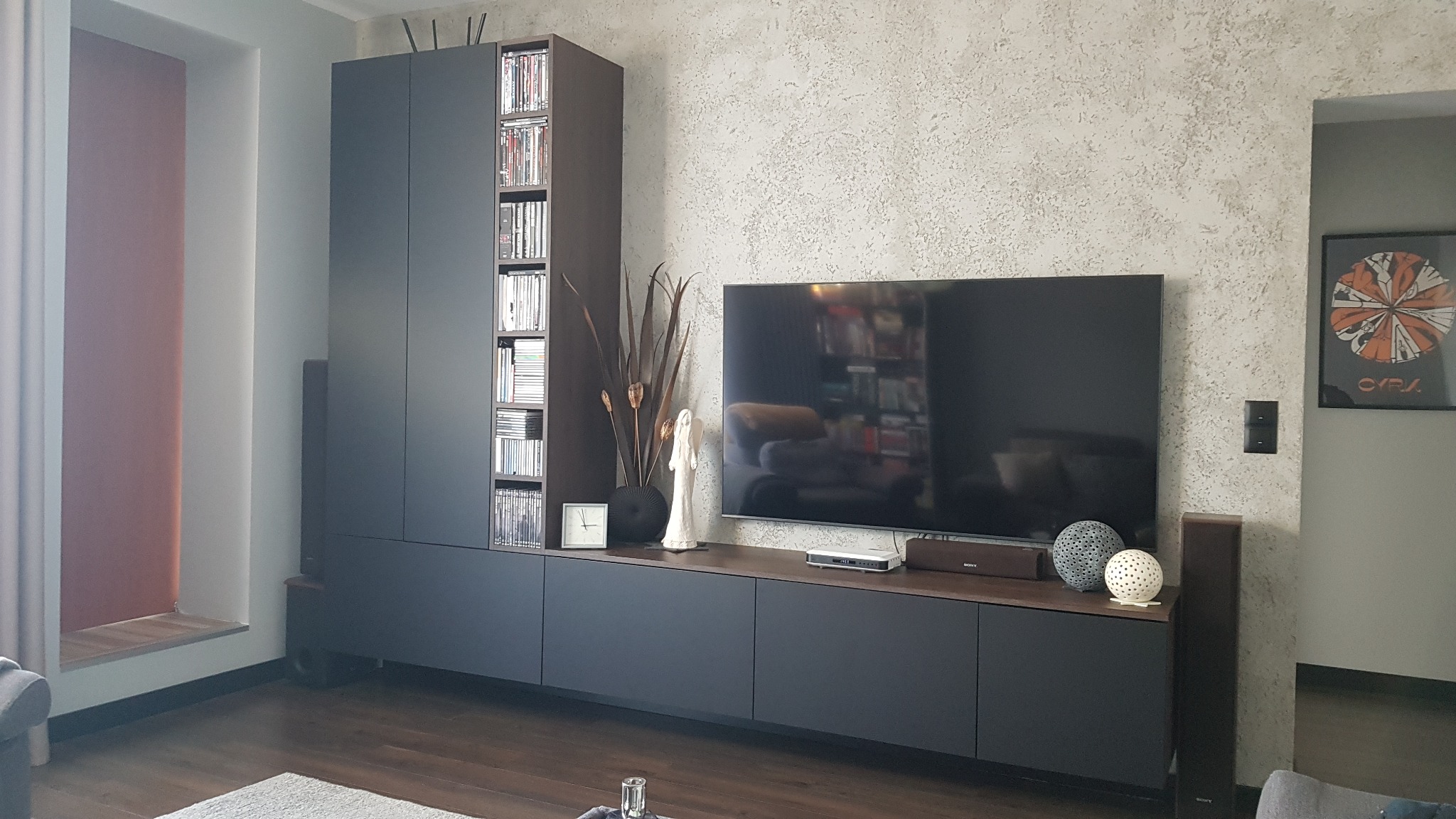 Nowoczesny salon z zabudową meblową w kolorze grafitowym, obejmującą szafę z półkami na płyty, szafkę RTV oraz dekoracje: zegar, figurka anioła i suszone trawy. Na ścianie widoczna faktura...