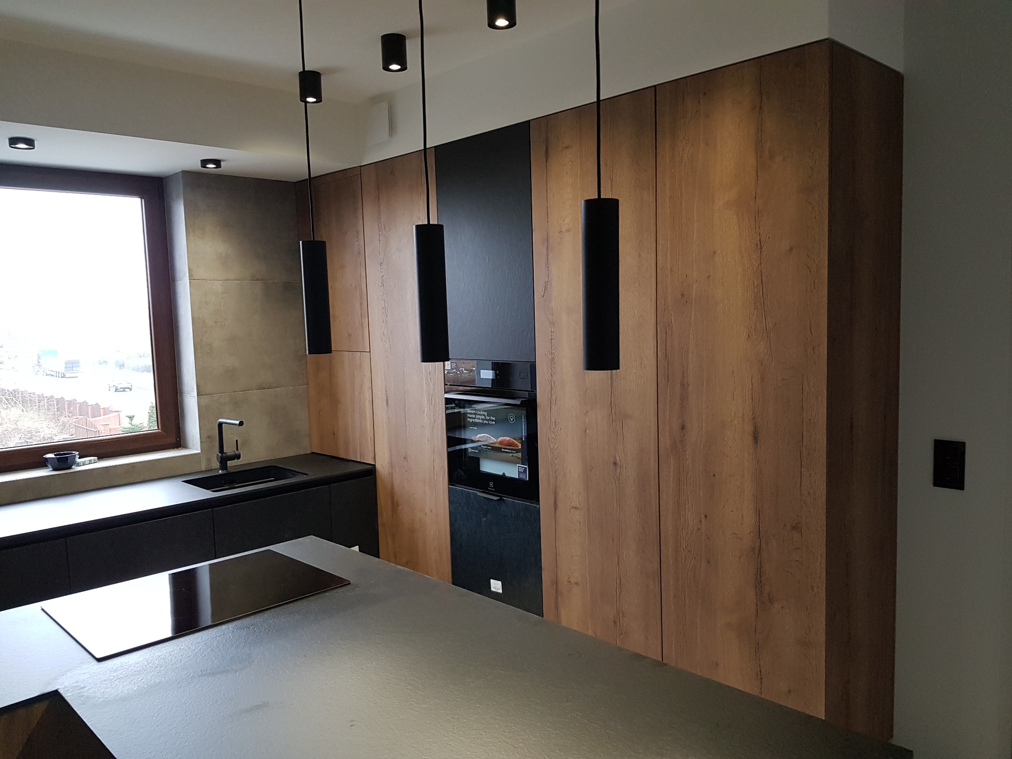 Nowoczesna kuchnia z drewnianymi frontami szafek, czarnym blatem i wiszącymi lampami cylindrycznymi, widok na okno z zewnątrz.