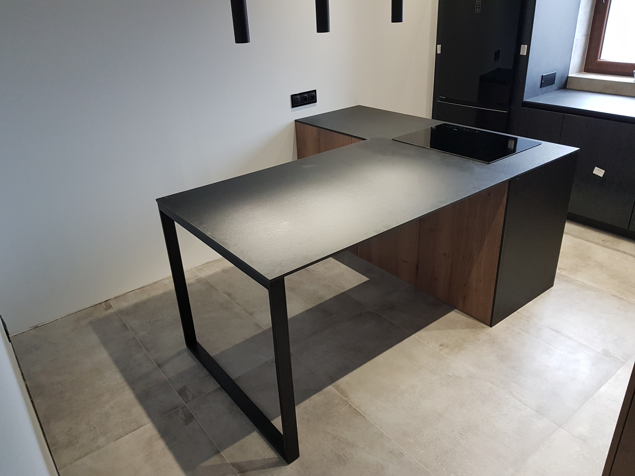 Nowoczesna kuchnia z wyspą o czarnym blacie i drewnianym wykończeniu, minimalistyczne czarne lampy wiszące, indukcyjna płyta grzewcza, widoczne gniazdka elektryczne na ścianie.