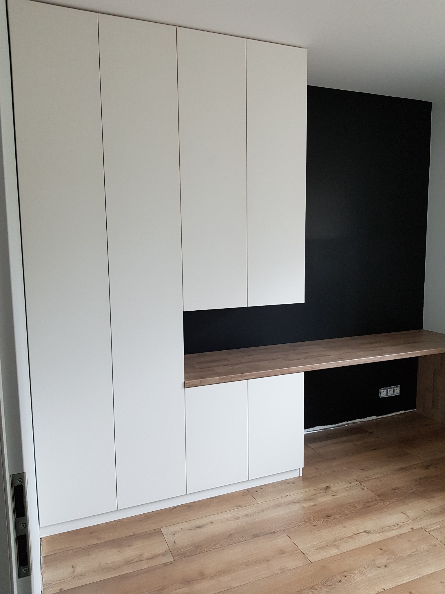 Biała zabudowa meblowa z blatem roboczym w kolorze drewna, na tle czarnej ściany, widoczne gniazdka elektryczne.