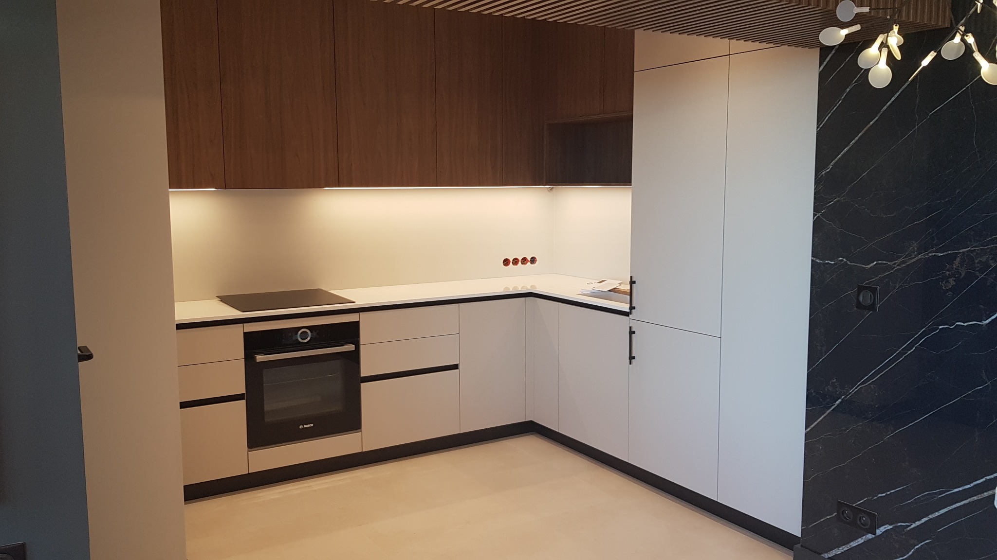 Nowoczesna kuchnia z białymi szafkami dolnymi z czarnymi detalami, drewnianymi szafkami górnymi i czarną ścianą z marmurowym wzorem oraz designerską lampą.