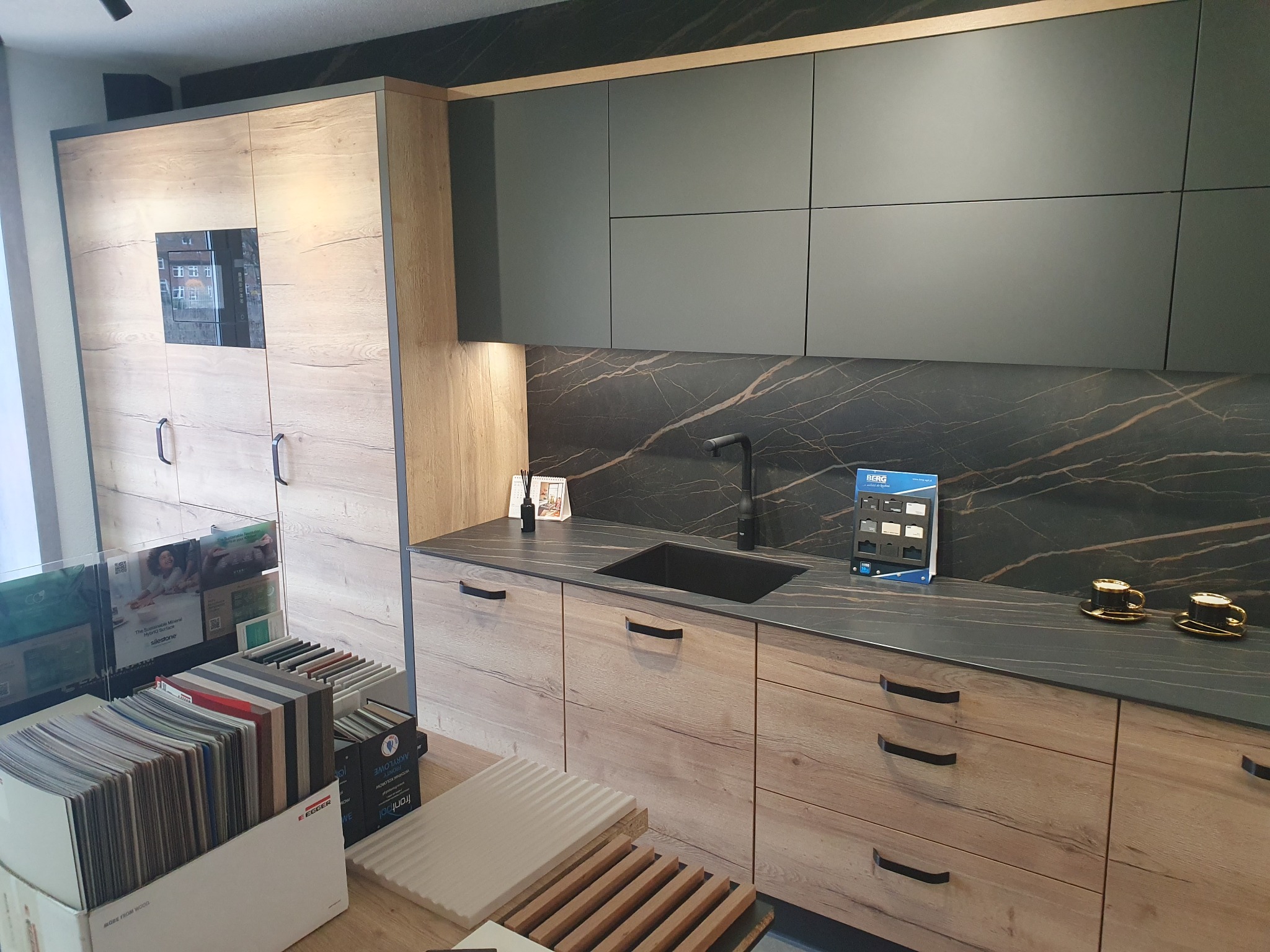 Ekspozycja nowoczesnej kuchni z drewnianymi frontami szafek dolnych i szarymi szafkami górnymi, czarny blat z marmurowym wzorem, zlewozmywak w kolorze czarnym, ekspozytory z próbkami materiałów...