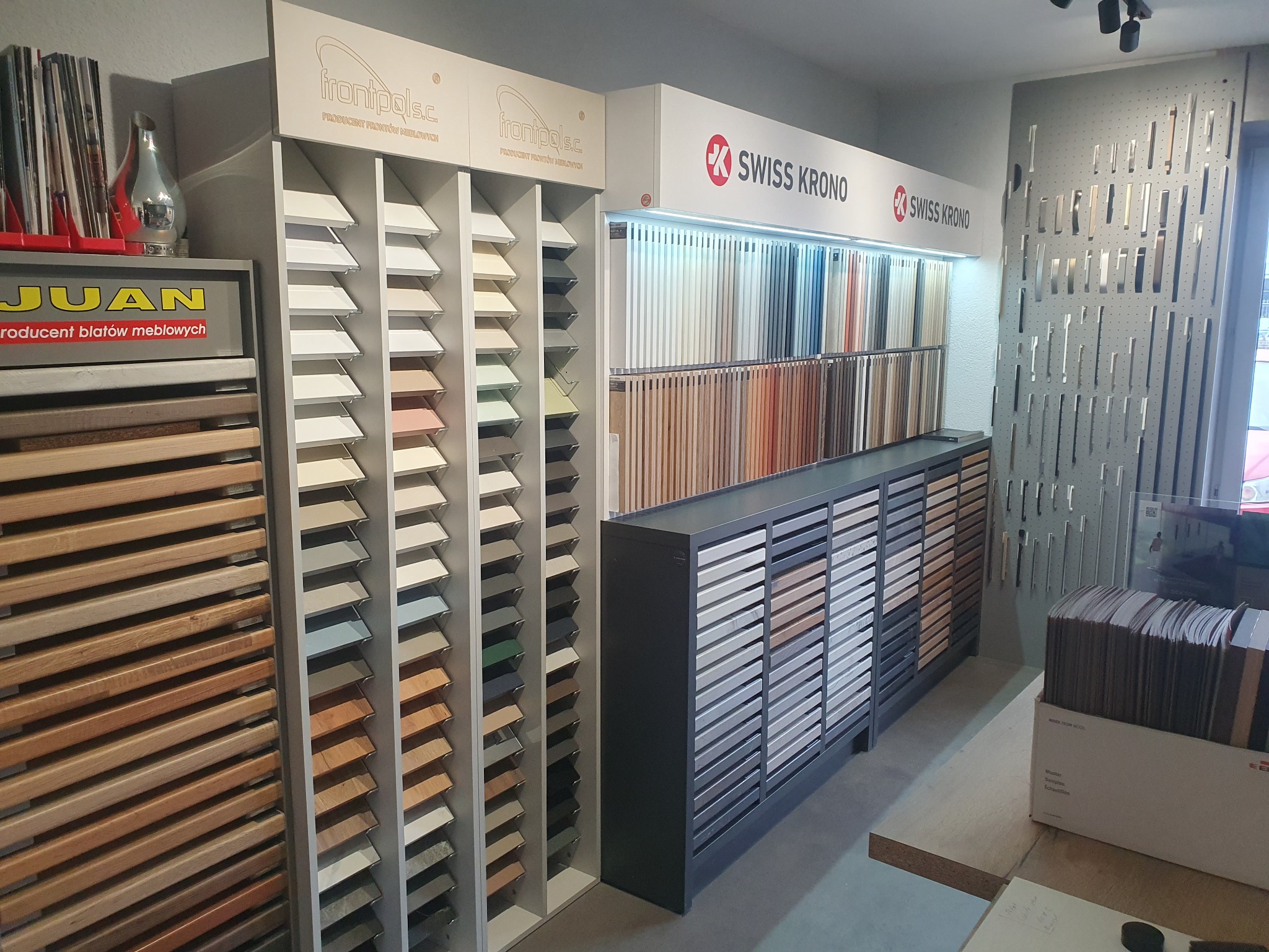 Ekspozycja próbek frontów meblowych i blatów w salonie, z logo producentów Frontpol i Swiss Krono, różne kolory i faktury drewna.