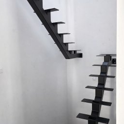 G-Steel GRZEGORZ ŚLĄCZKA - Nowoczesne, czarne schody metalowe wspornikowe, zamocowane do białej ściany. Widoczne betonowe schody poniżej. Minimalistyczny design.