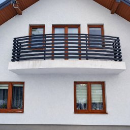 G-Steel GRZEGORZ ŚLĄCZKA - Biały dom z balkonem i czarną, poziomą balustradą. Trzy okna na balkonie, dwa poniżej. Drewniane ramy okienne i brązowe wykończenie dachu. Minimalistyczny design.