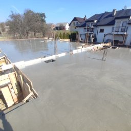 BOHDAN LUKIANIV - Świeżo wylany beton na przygotowanym fundamencie z wystającymi prętami zbrojeniowymi i drewnianym szalunkiem, w tle zabudowa jednorodzinna w słoneczny dzień.
