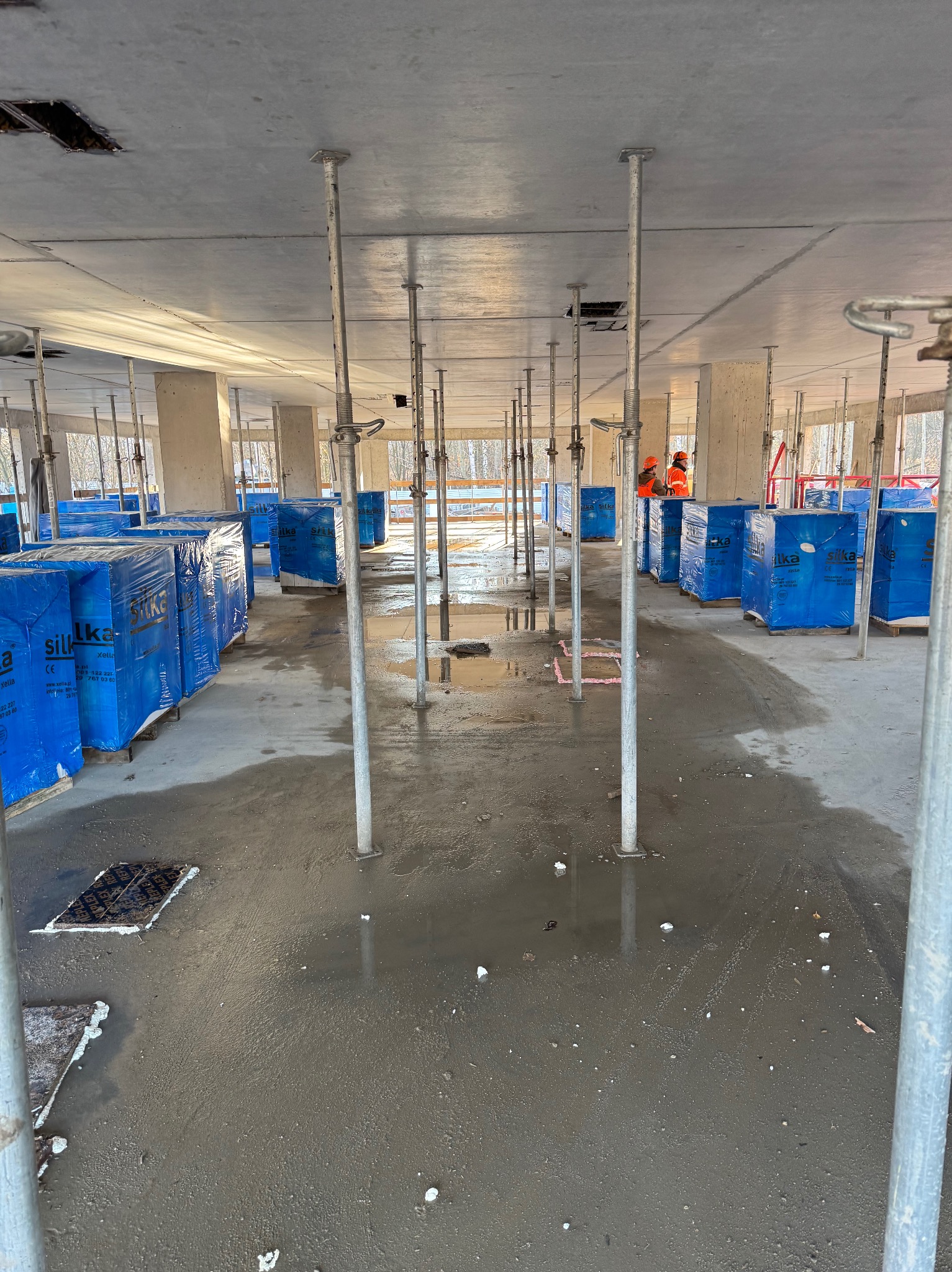 Wnętrze budynku w trakcie budowy, widoczne podpory stropowe, mokra betonowa posadzka i palety z niebieskimi bloczkami Silka Xella.