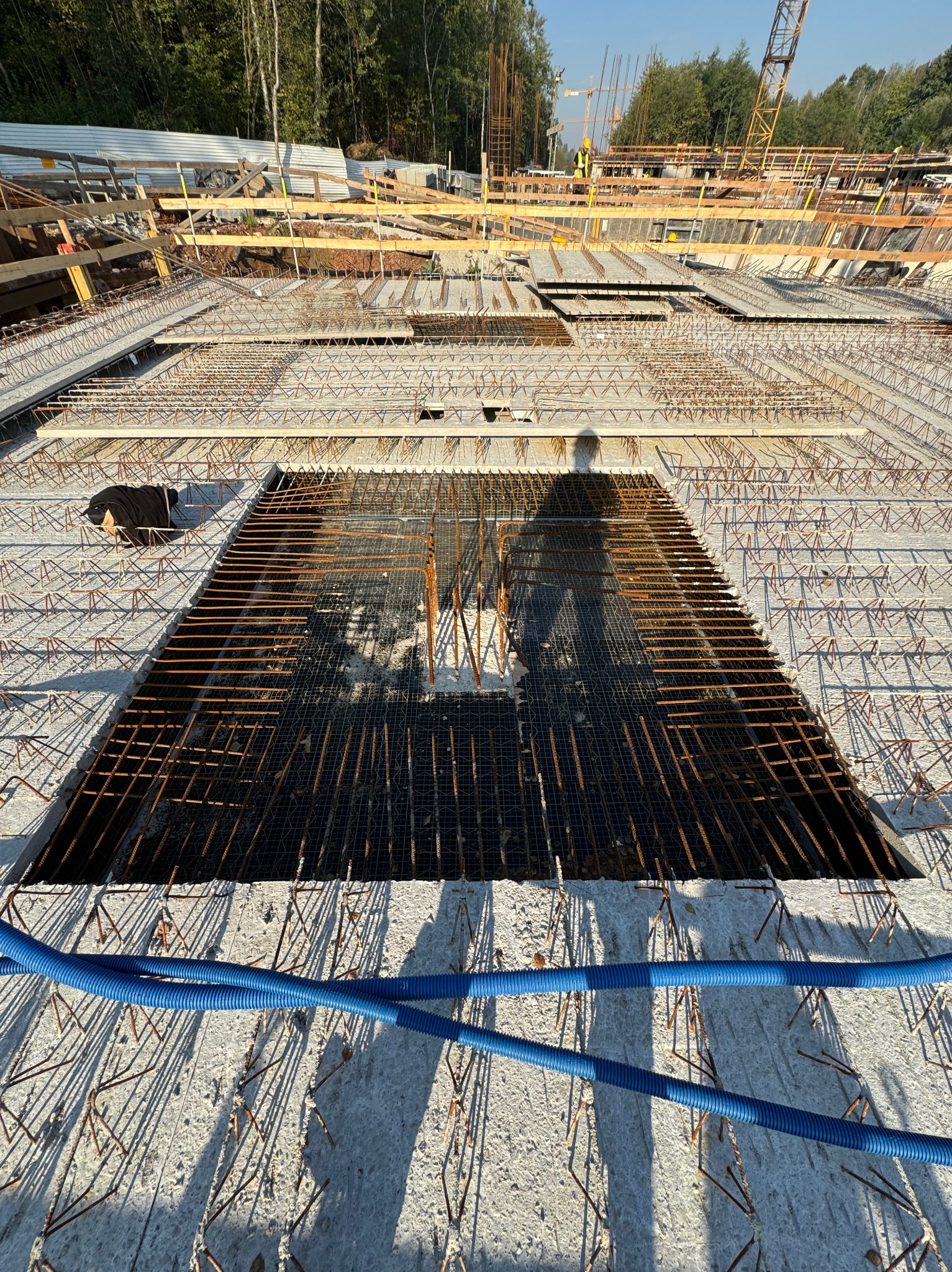 Zbrojenie stropu z otworem, widoczne niebieskie rury instalacyjne i dalszy plan budowy z dźwigami.