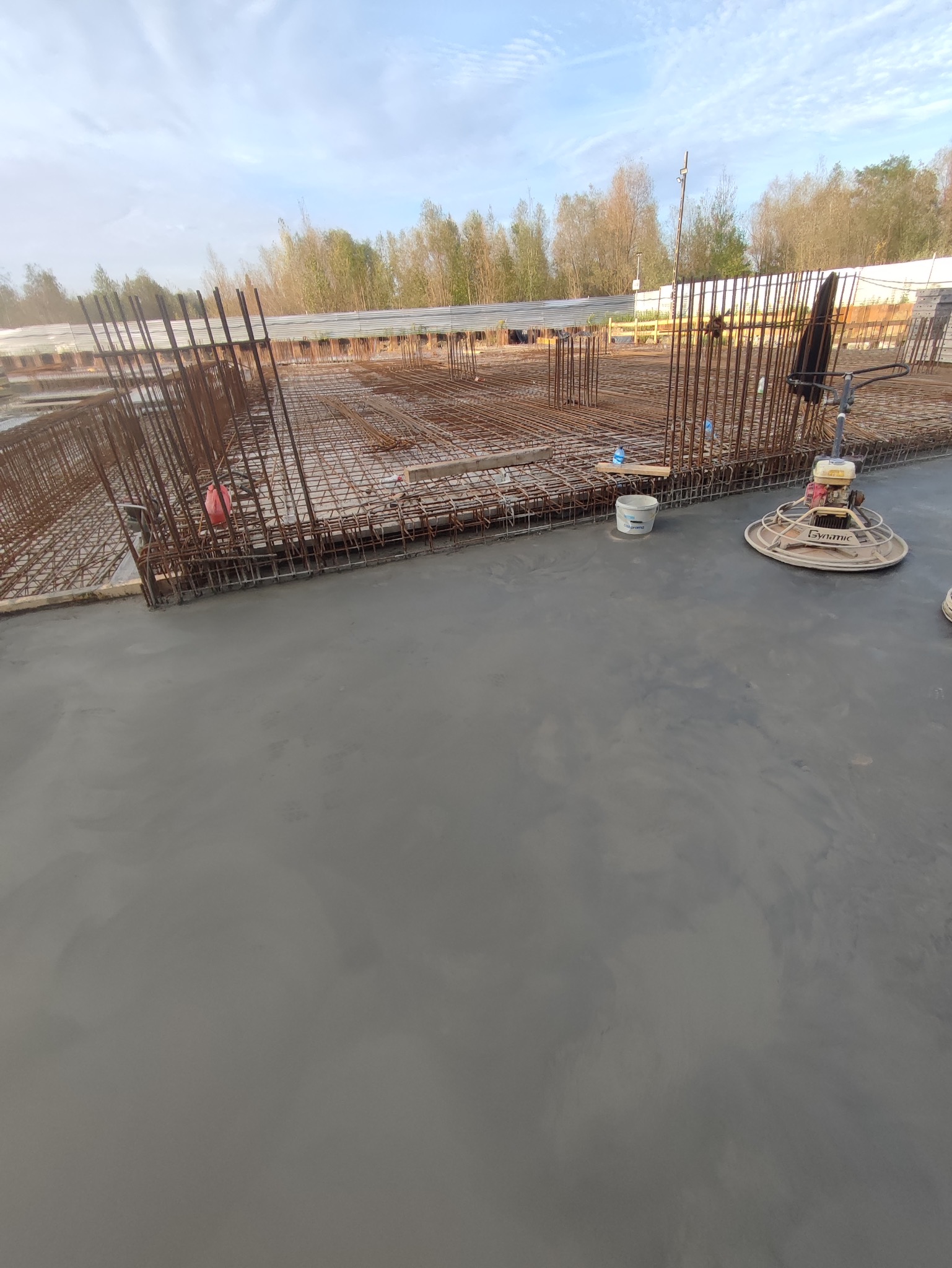 Wygładzona, szara betonowa posadzka z widocznym zbrojeniem i maszyną do zacierania betonu Dynamic w tle, na budowie z drzewami w tle.