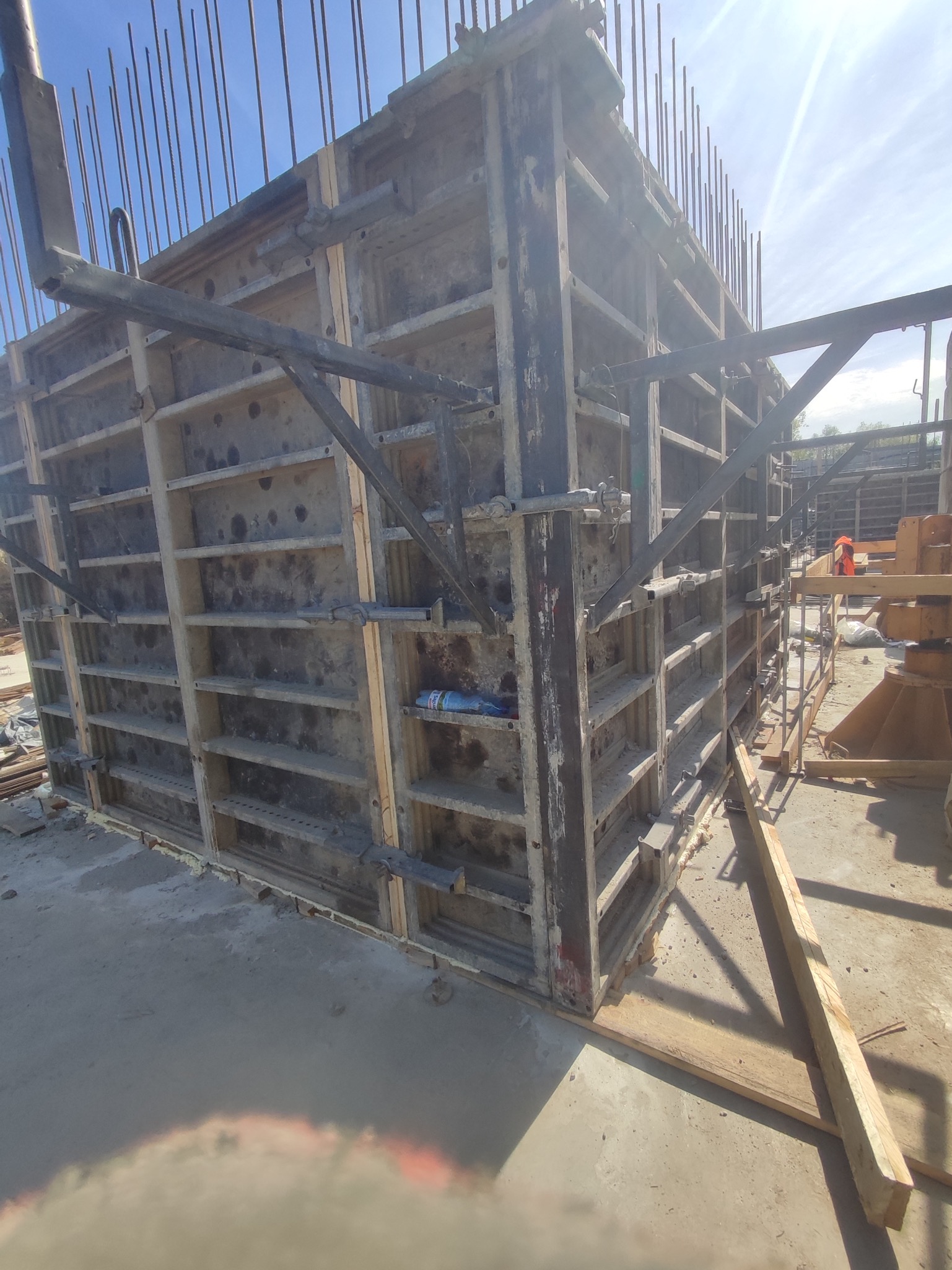 Szare szalunki żelbetowe z wystającymi prętami zbrojeniowymi, wzmocnione metalowymi wspornikami, widoczne elementy drewniane i butelka wody wewnątrz konstrukcji.