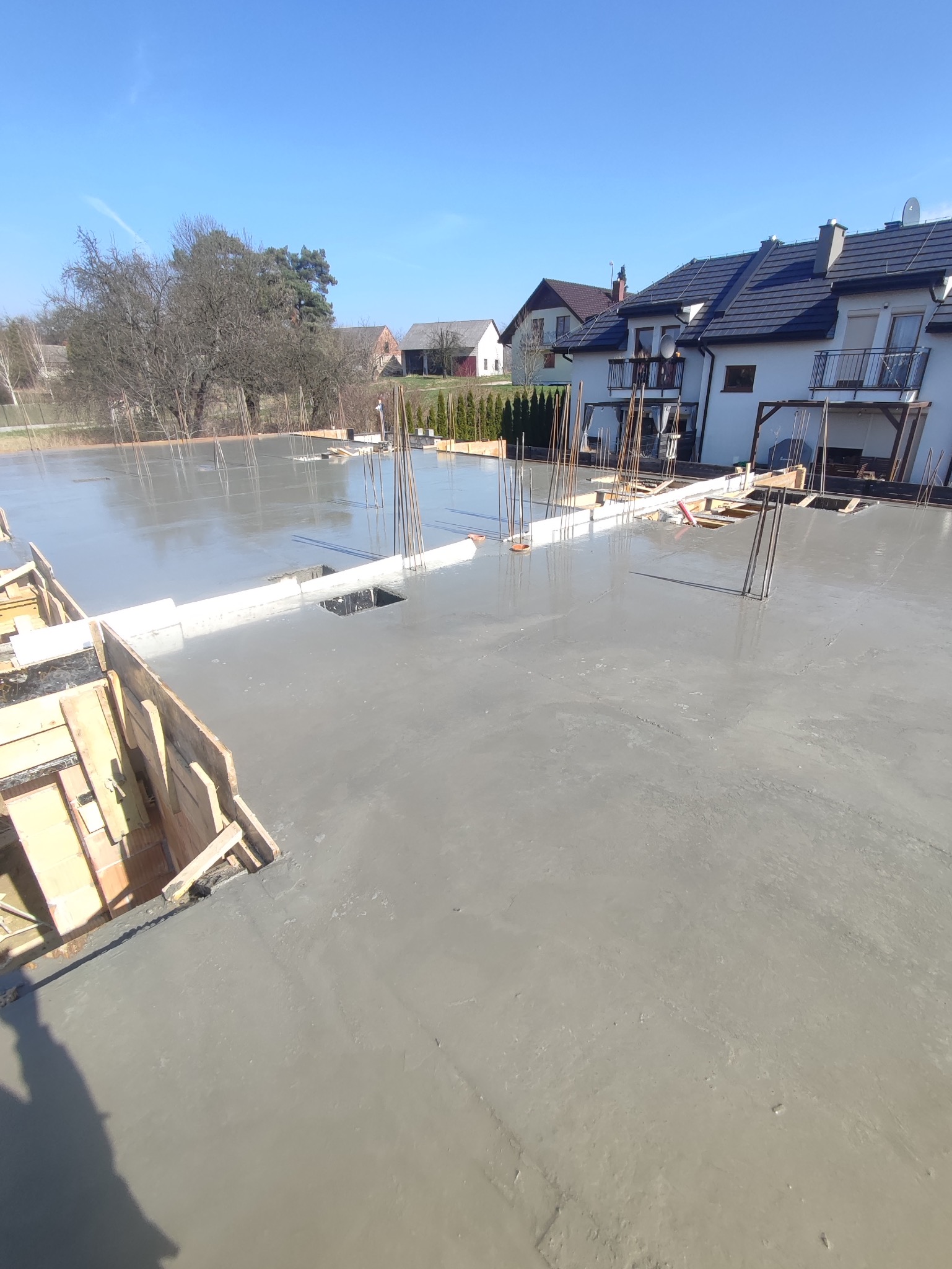 Świeżo wylany beton na przygotowanym fundamencie z wystającymi prętami zbrojeniowymi i drewnianym szalunkiem, w tle zabudowa jednorodzinna w słoneczny dzień.