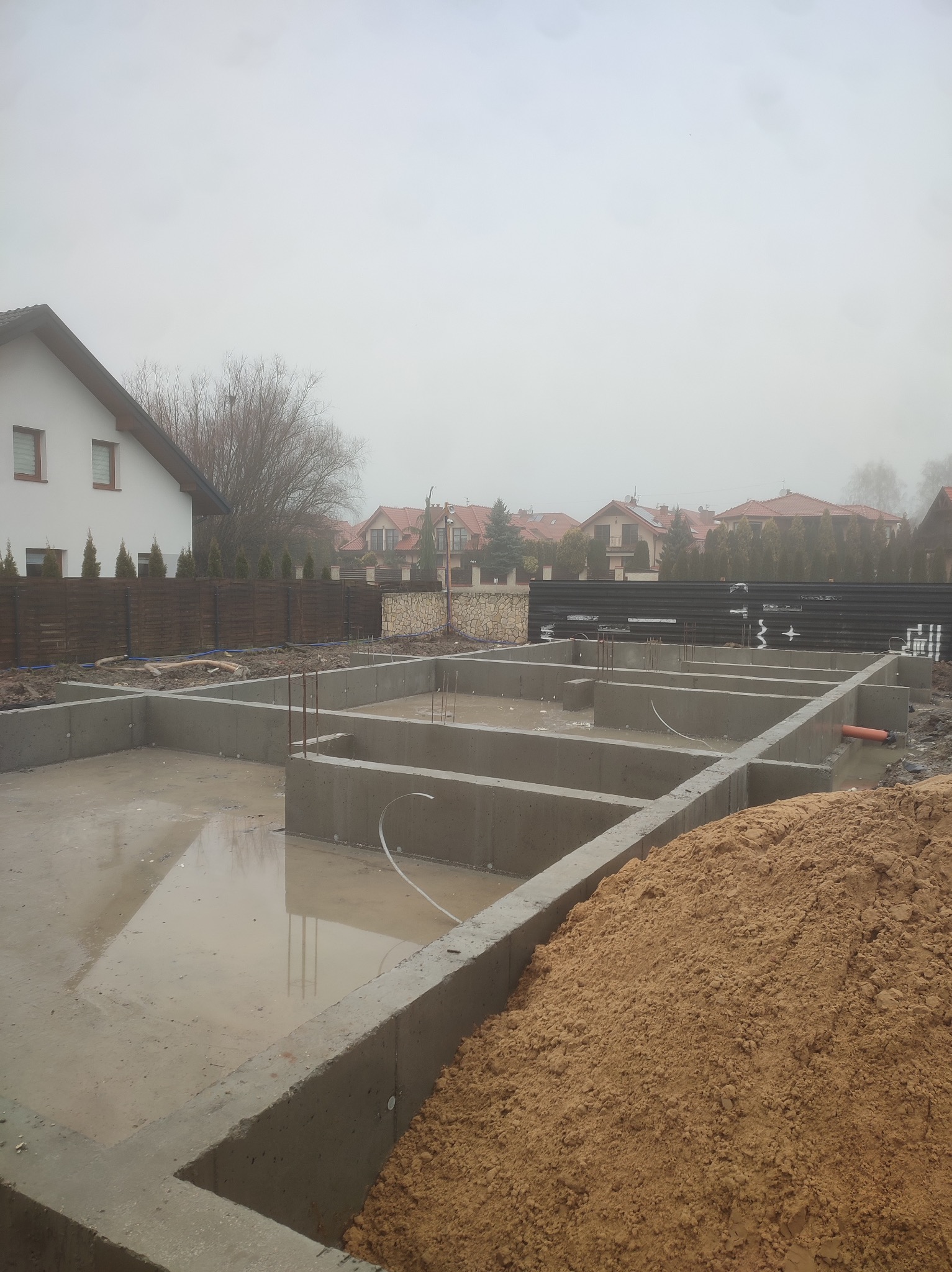 Zalane wodą betonowe fundamenty domu jednorodzinnego w trakcie budowy, widoczny stos piasku i zabudowa sąsiedzka w tle.