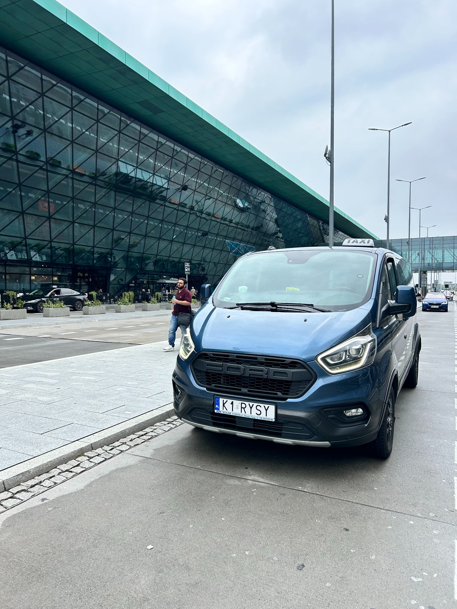 Niebieski bus Ford Transit z napisem TAXI na dachu, zaparkowany przed nowoczesnym budynkiem lotniska z przeszkloną fasadą i zielonym dachem. Widoczny mężczyzna w oddali.
