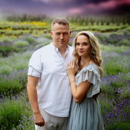 Fotobambino Anna Gąsiorowska - Para w objęciach na polu lawendy z dramatycznym niebem w tle, kobieta w zwiewnej sukience, mężczyzna w białej koszuli i krótkich spodenkach.