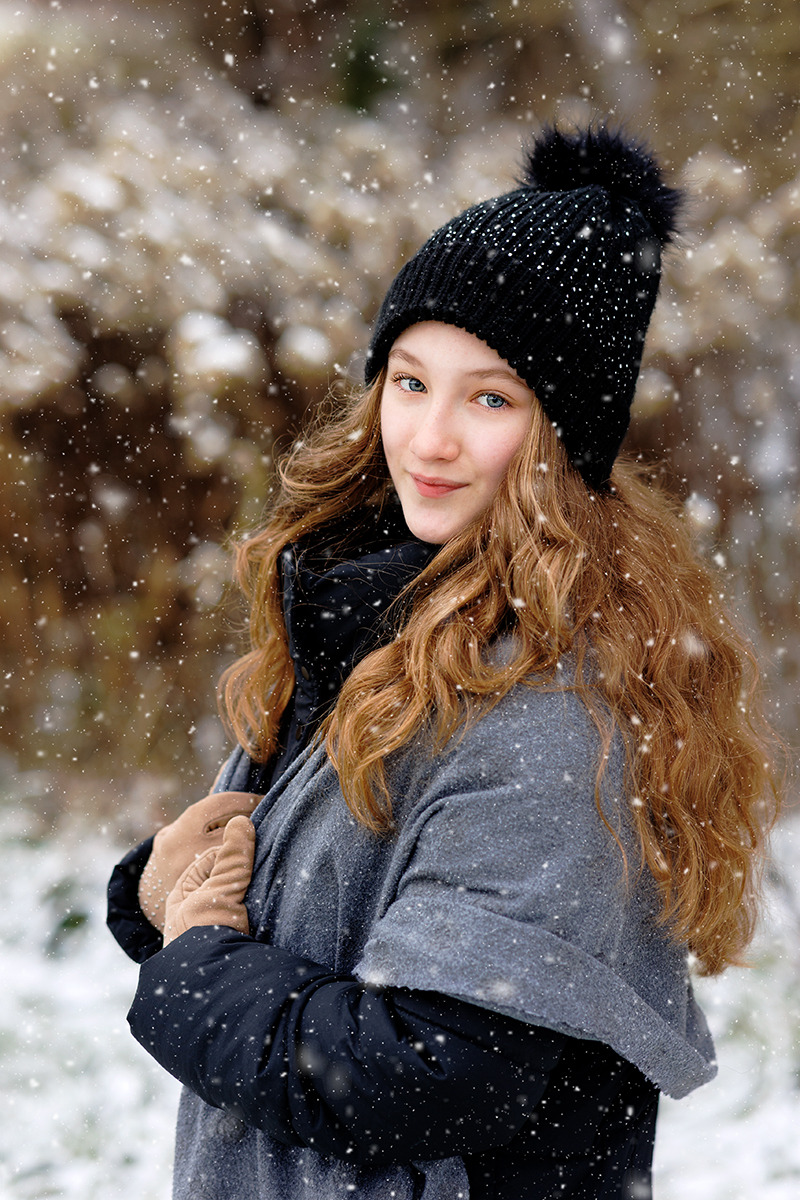 Portret uśmiechniętej nastolatki w czapce z pomponem i szarym płaszczu na tle zimowego krajobrazu podczas opadów śniegu.