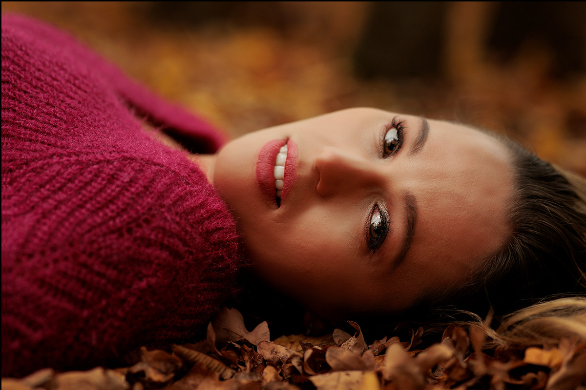 Portret kobiety leżącej na opadłych liściach w jesiennej scenerii, ubrana w bordowy sweter, z delikatnym uśmiechem i błyszczącym makijażem oczu.