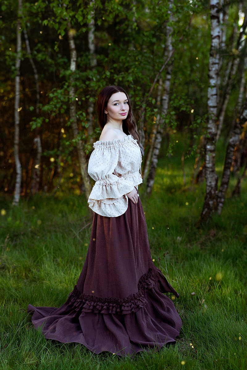 Portret kobiety w zwiewnej bluzce z falbanami i długiej spódnicy w leśnej scenerii z brzozami i delikatnym oświetleniem.