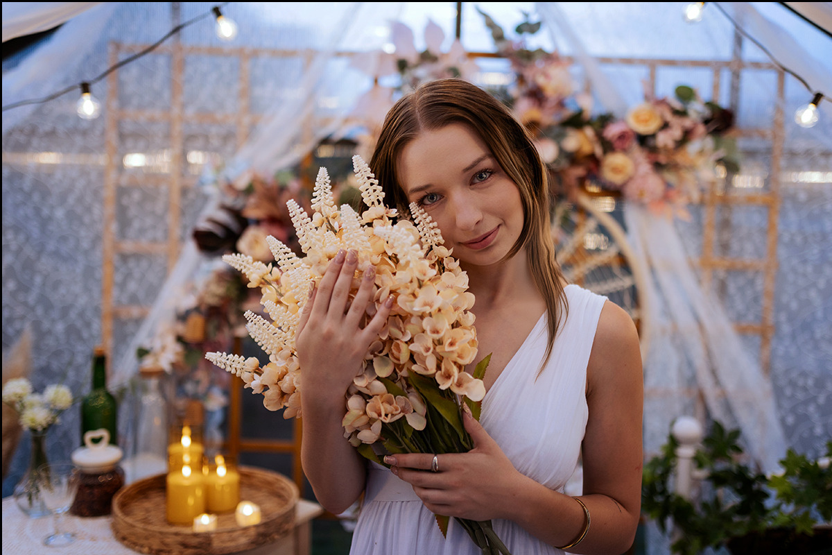 Uśmiechnięta kobieta w białej sukience trzyma kremowy bukiet kwiatów na tle dekoracji ślubnych z lampkami, świecami i kwiatami w rustykalnym stylu.
