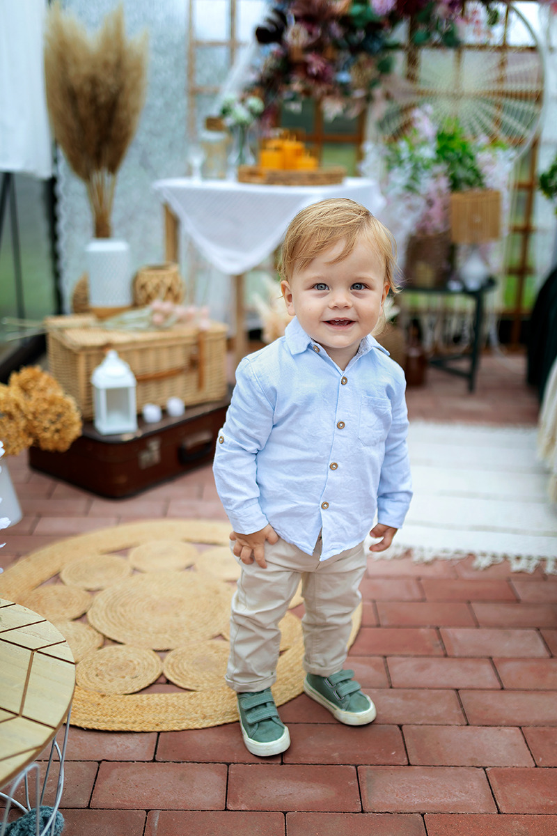 Uśmiechnięty roczny chłopiec w błękitnej koszuli i beżowych spodniach, stojący na ceglanej podłodze z jutowym dywanem, w tle kompozycja kwiatowa i dekoracje rustykalne.
