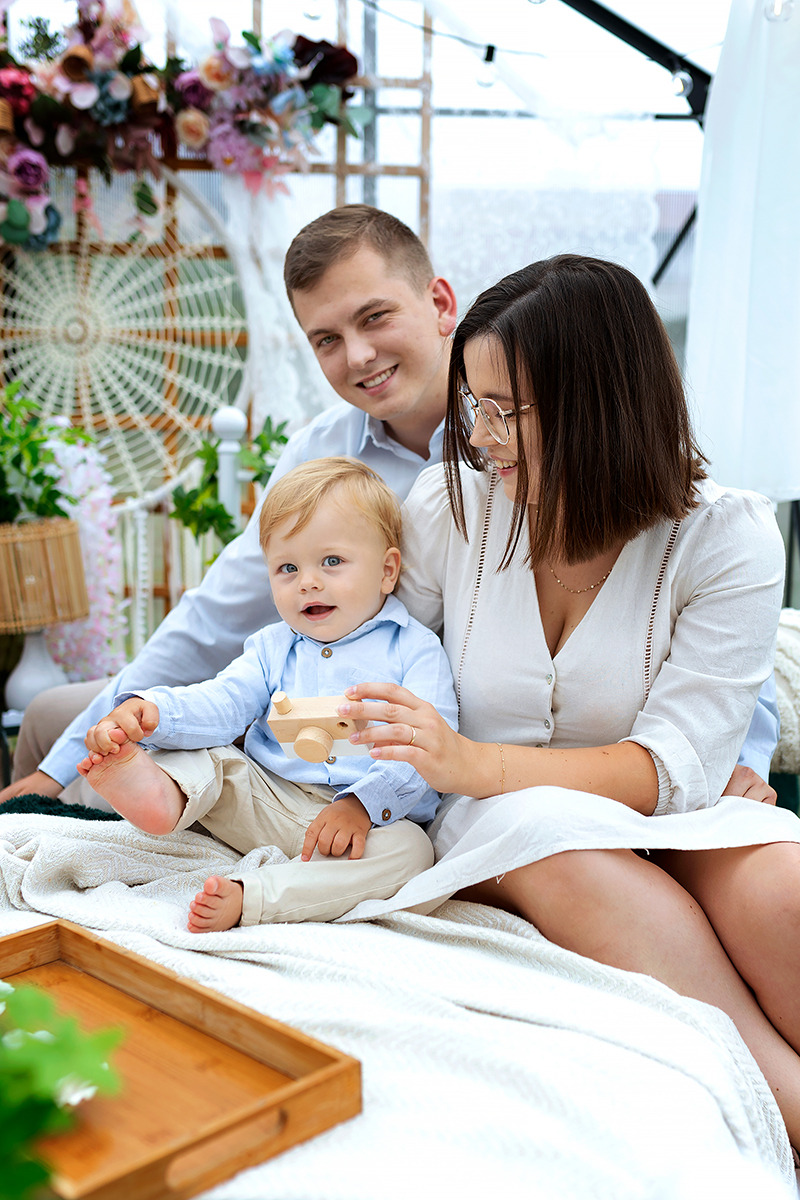 Rodzina: uśmiechnięty ojciec, matka w okularach i mały chłopiec z drewnianym aparatem siedzą na łóżku w jasnym pomieszczeniu z kwiatową dekoracją w tle.