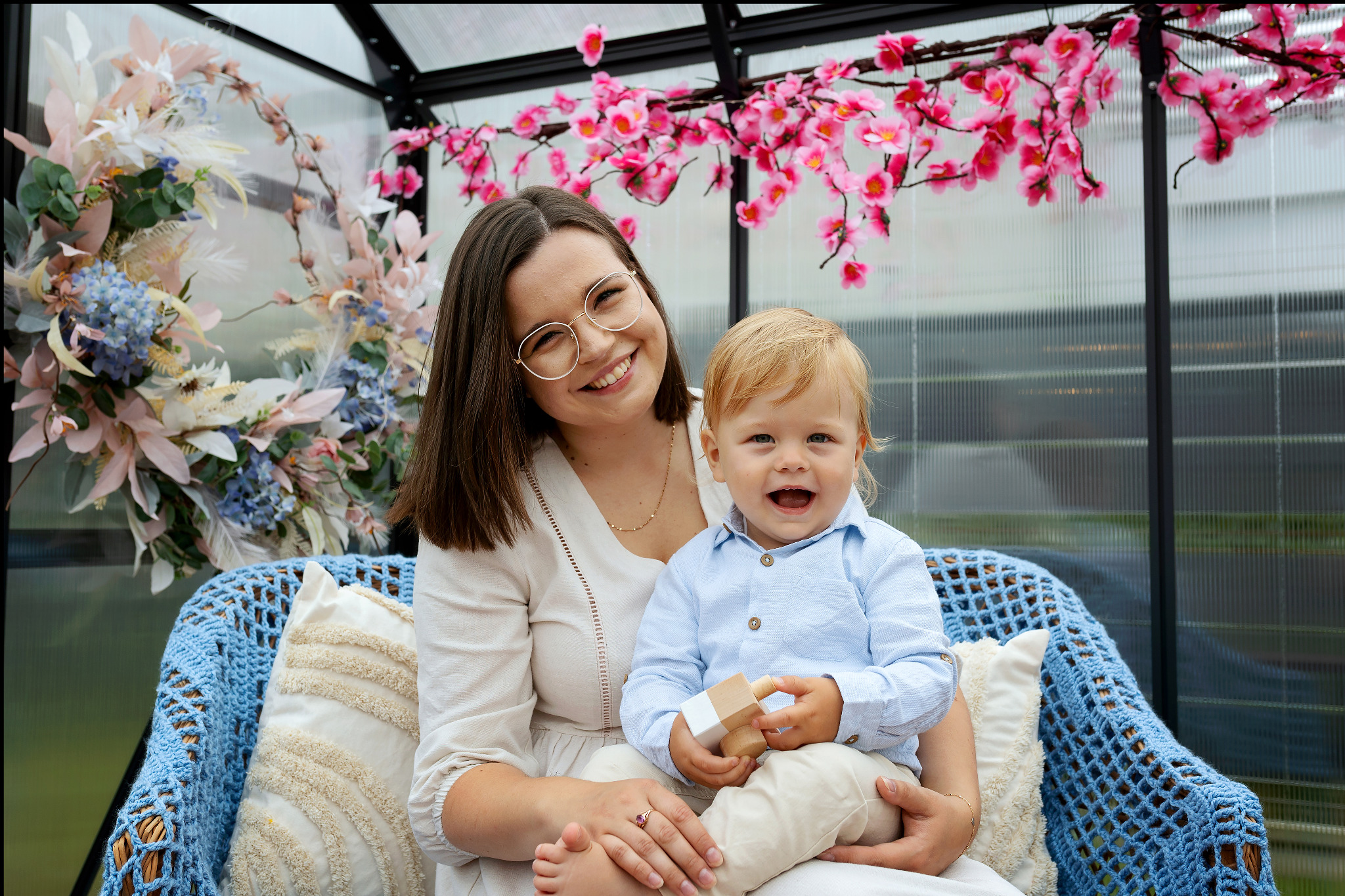 Uśmiechnięta mama w okularach trzyma na kolanach roześmianego synka w fotelu z niebieskiego rattanu na tle dekoracji kwiatowych w Bolesławcu.