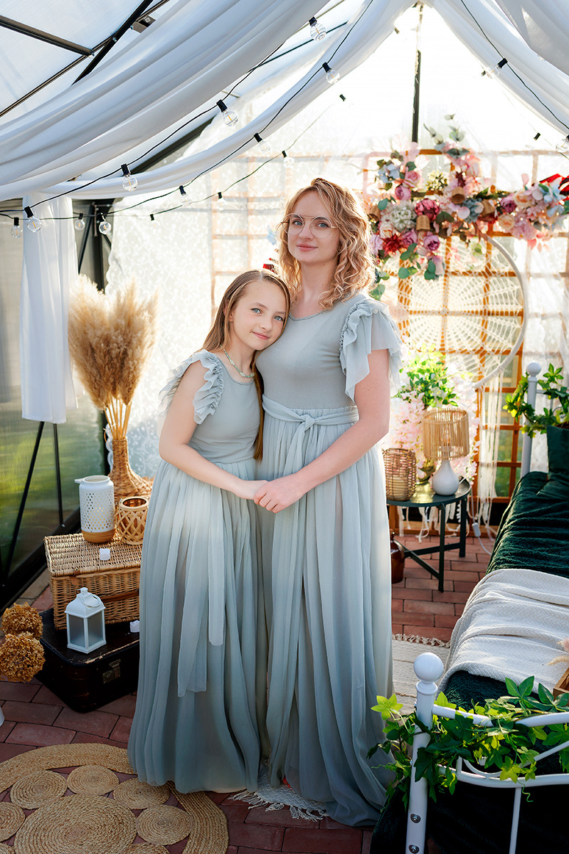 Matka i córka w dopasowanych, zwiewnych, szarozielonych sukienkach pozują w dekoracyjnej szklarni z kwiatami i lampionami.
