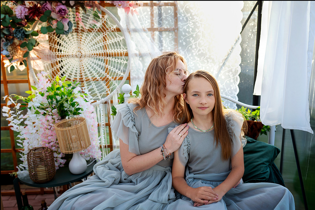 Matka całuje córkę w czoło podczas sesji zdjęciowej w plenerze, obie ubrane w identyczne, zwiewne sukienki w odcieniu szaro-zielonym, na tle dekoracji z kwiatów i łapacza snów.