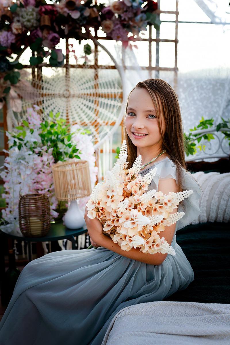 Młoda dziewczyna w zwiewnej, szarej sukience siedzi na łóżku, trzymając bukiet sztucznych kwiatów w odcieniach bieli i brzoskwini, z uśmiechem patrzy w obiektyw, tło stanowi dekoracja z kwiatów...