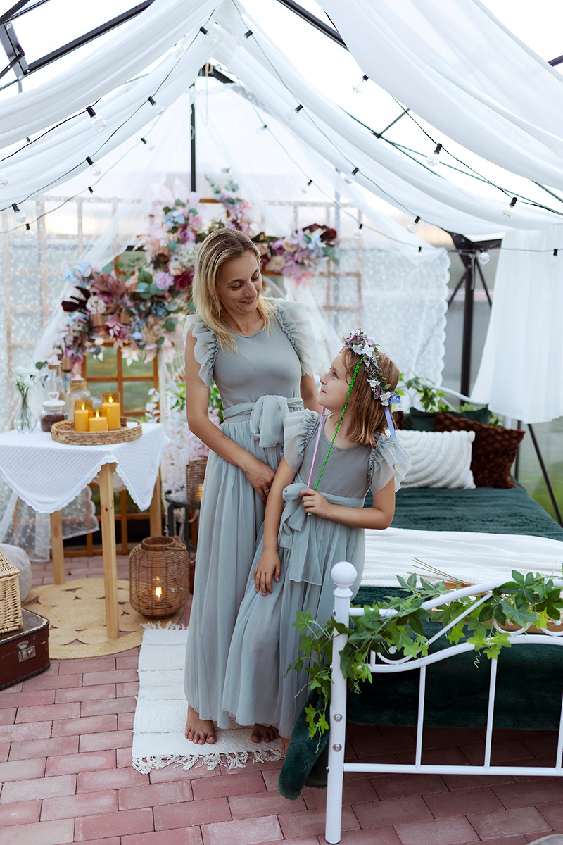 Matka i córka w dopasowanych, szarozielonych sukienkach pozują w dekoracyjnej scenerii z baldachimem, kwiatami i świecami, tworząc ciepłą i rodzinną atmosferę.