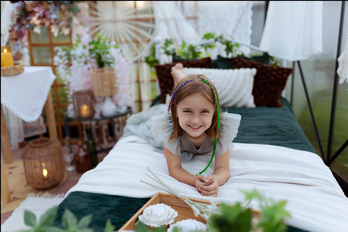 Uśmiechnięta dziewczynka leży na łóżku w dekoracyjnej scenerii z kwiatami i lampionami, włosy ozdobione kolorowymi warkoczykami.