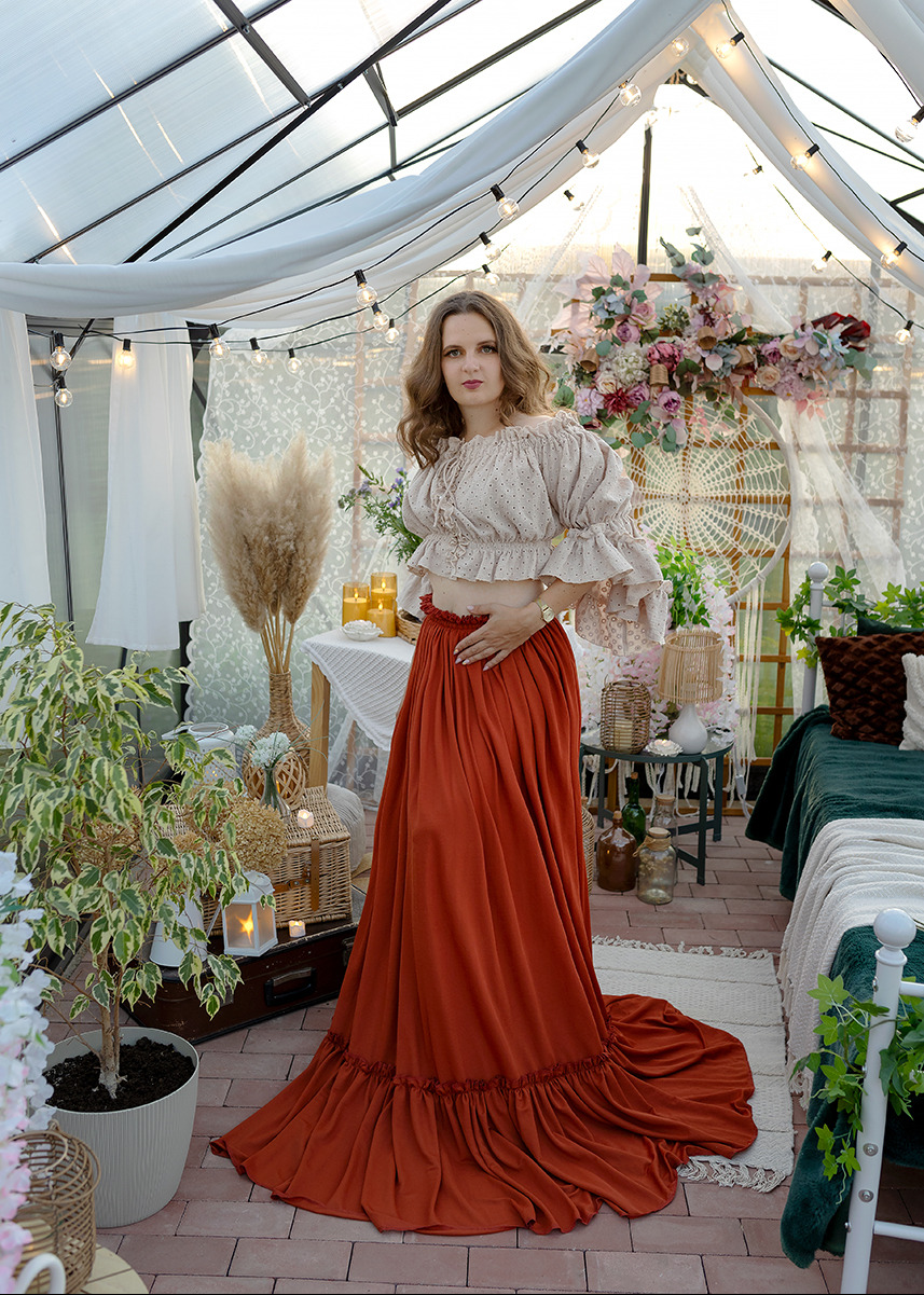 Kobieta w rustykalnym stroju pozuje w szklarni z dekoracjami boho, oświetlona girlandami żarówek, z widocznymi elementami wystroju jak łapacze snów i kwiatowe kompozycje.