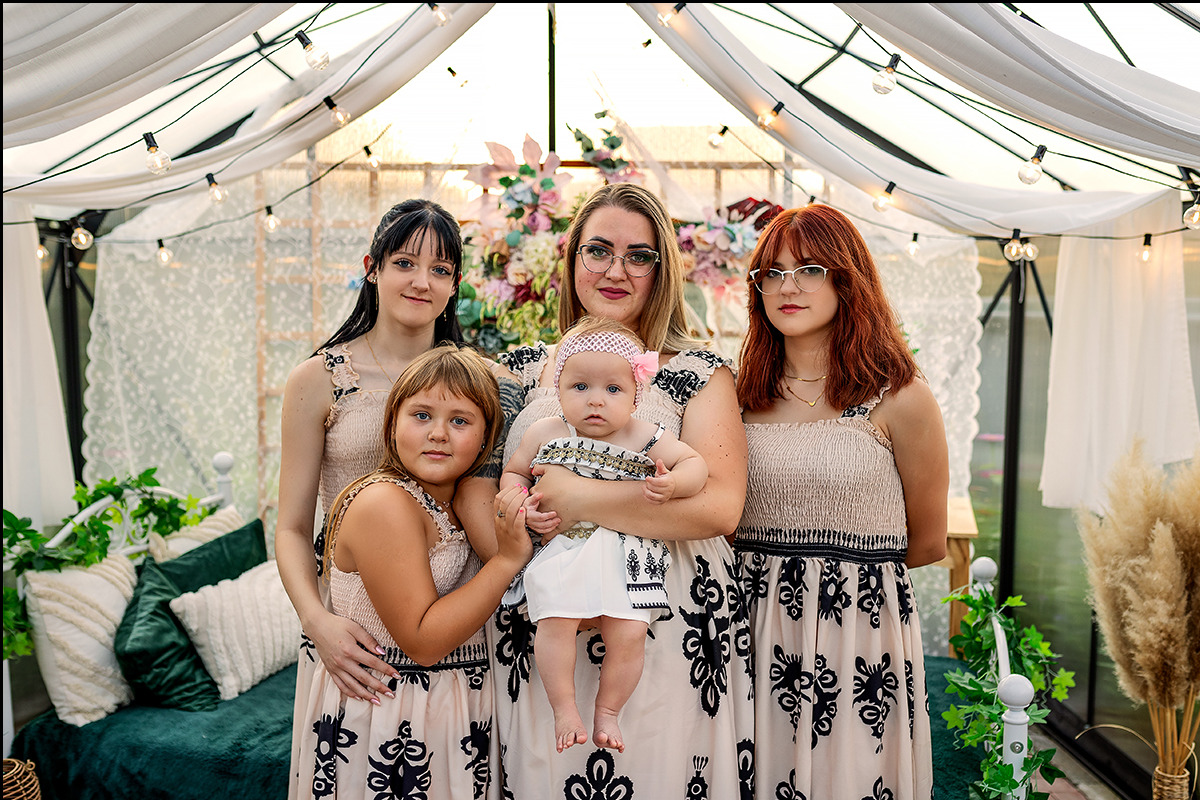 Portret rodzinny: mama trzyma niemowlę, obok trzy córki w pasujących sukienkach na tle altany z kwiatami i lampkami.