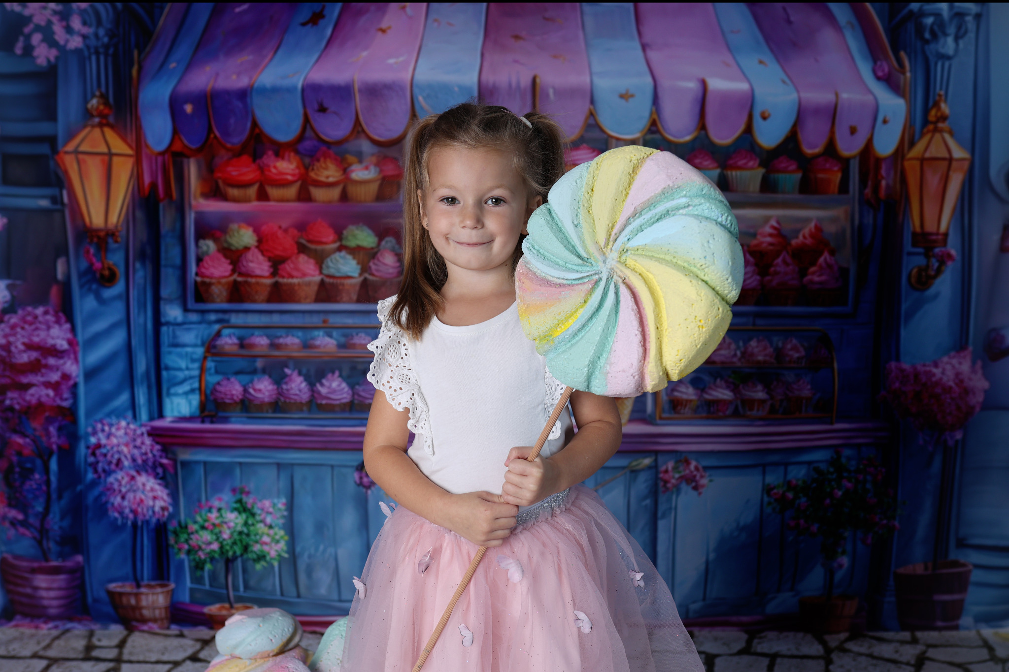 Uśmiechnięta dziewczynka w tiulowej spódniczce trzymająca duży, pastelowy lizak na tle malowanej scenerii cukierni z babeczkami.