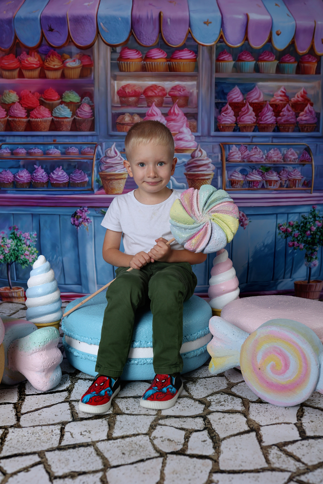 Chłopiec w białej koszulce i zielonych spodniach siedzi na makaroniku, trzymając lizaka na tle dekoracji cukierniczych i witryny sklepowej z babeczkami.