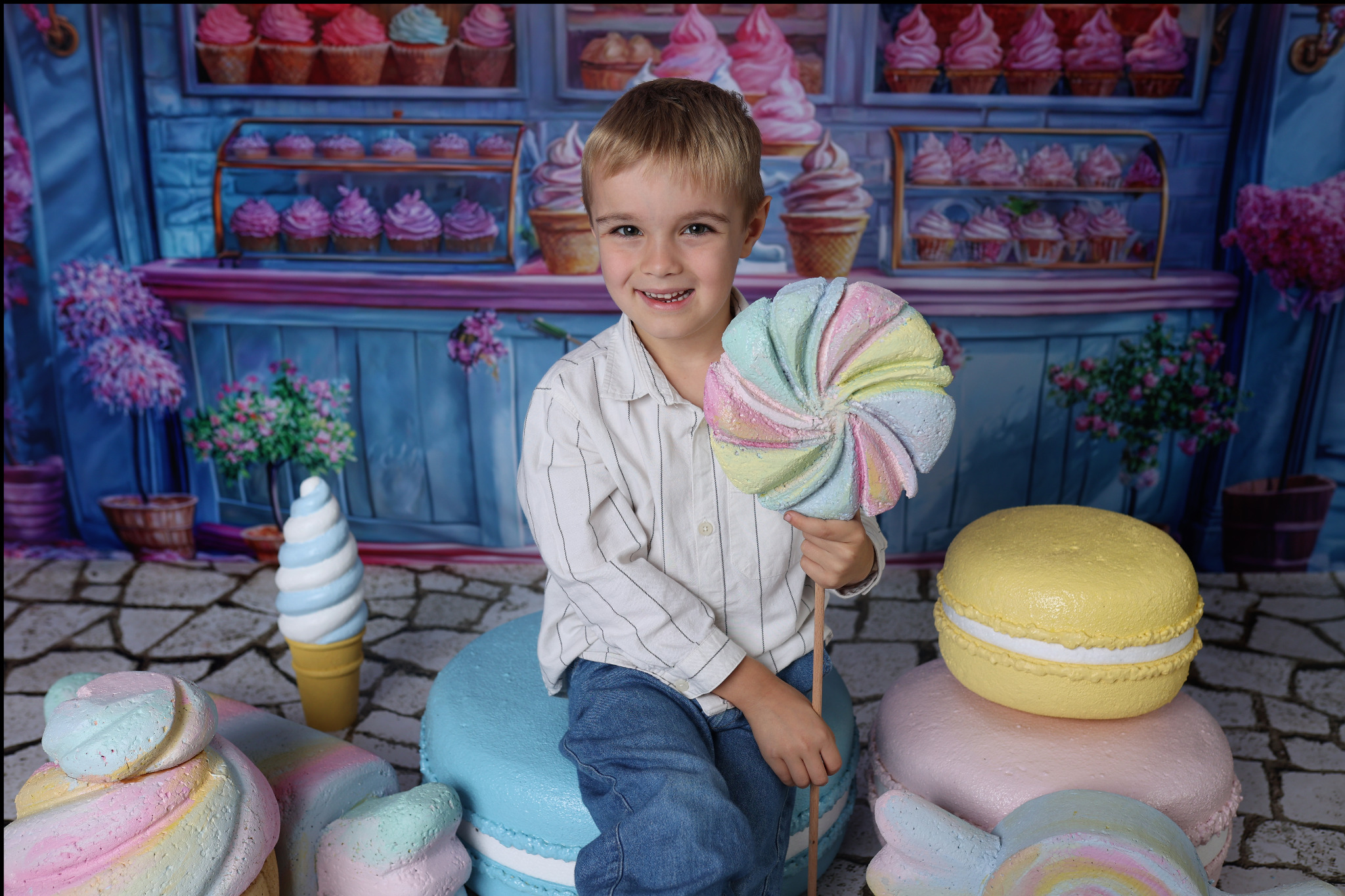 Uśmiechnięty chłopiec siedzi na makaroniku, trzymając w ręku lizaka z pastelowymi kolorami na tle dekoracji cukierniczej.