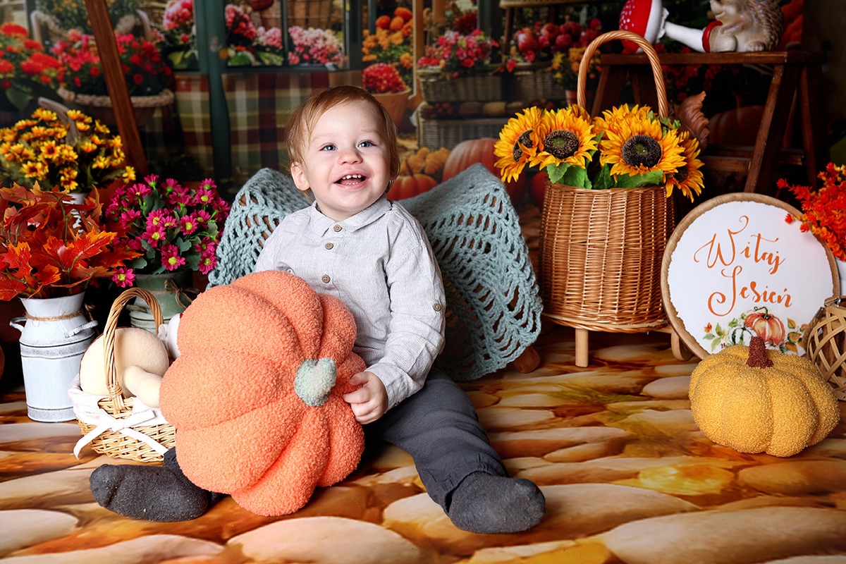 Uśmiechnięty chłopiec siedzi na podłodze, trzymając pluszową dynię na tle jesiennej scenerii z koszem słoneczników i dekoracją z napisem 'Witaj Jesieni'.